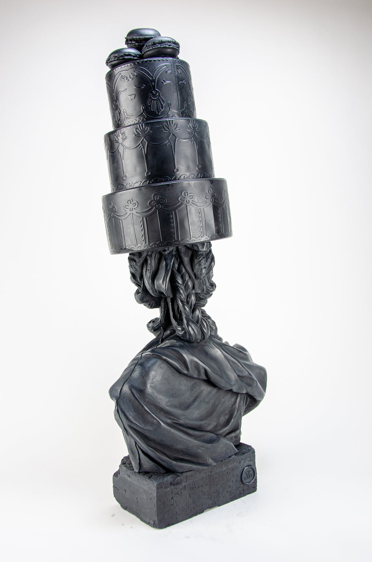 Tall dark charcoal sculpture of Marie Antoinette appearing from the back right wearing a giant three-layered cake with charcoal Macrons on top of her head. 