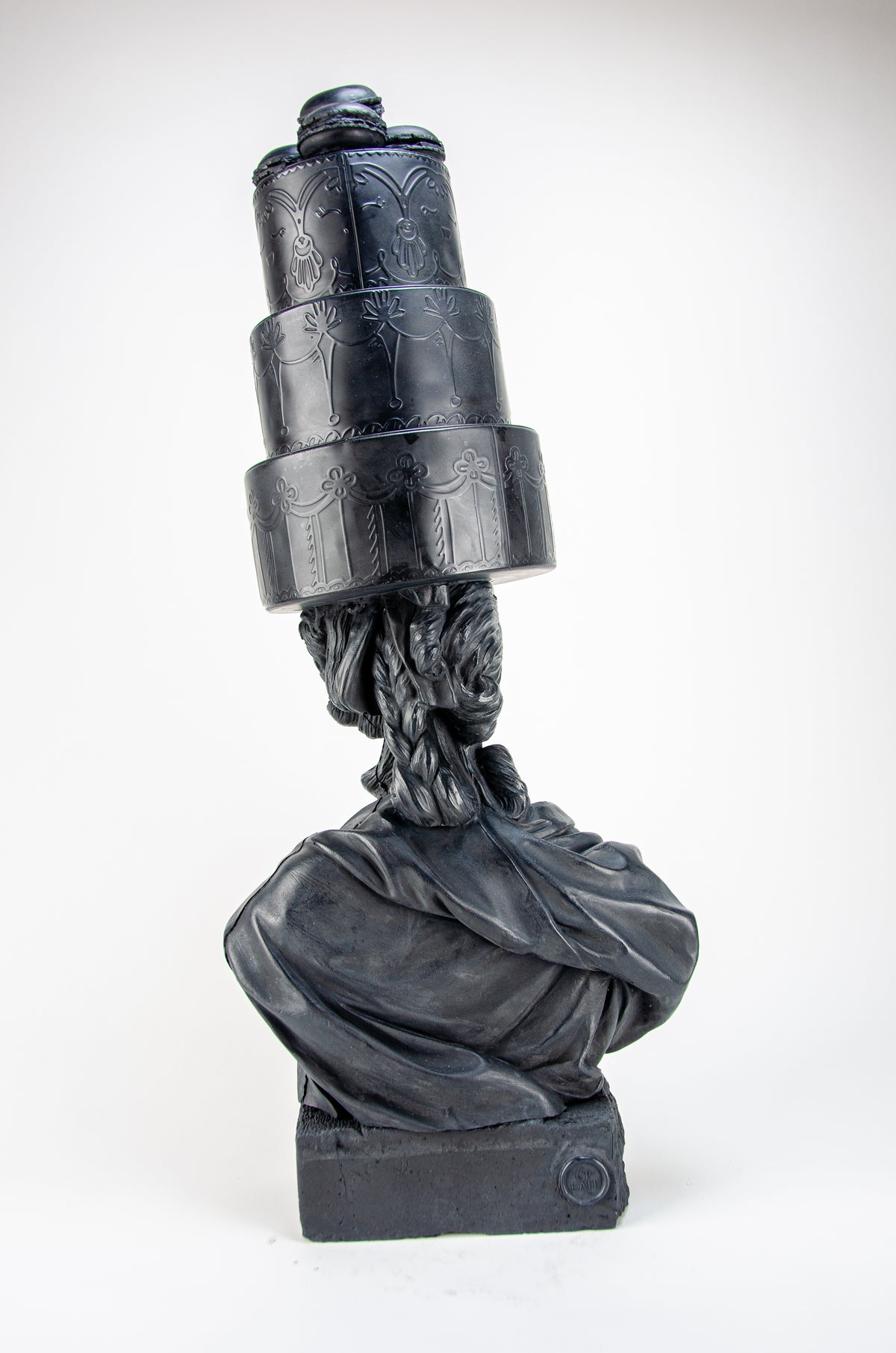 Tall dark charcoal sculpture of Marie Antoinette appearing from the back wearing a giant three-layered cake with charcoal Macrons on top of her head.  