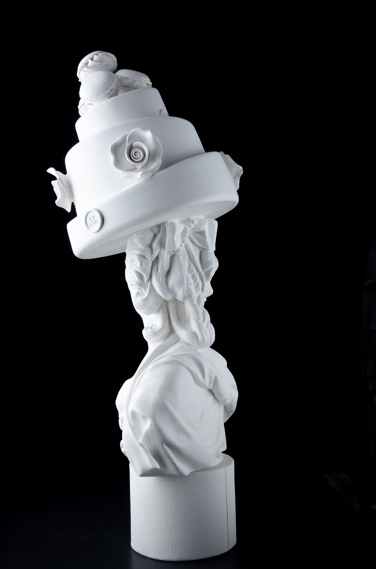 Tall bright white sculpture of Marie Antoinette looking backwards wearing a large whimsical cake with roses and Macron cookies on her head. Dark black background.