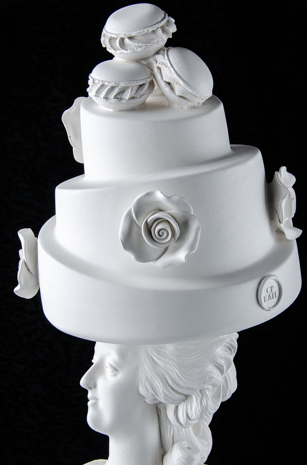White sculpture bust of Marie Antoinette looking to the side wearing a large whimsical cake with roses and Macron cookies on her head. Dark black background.