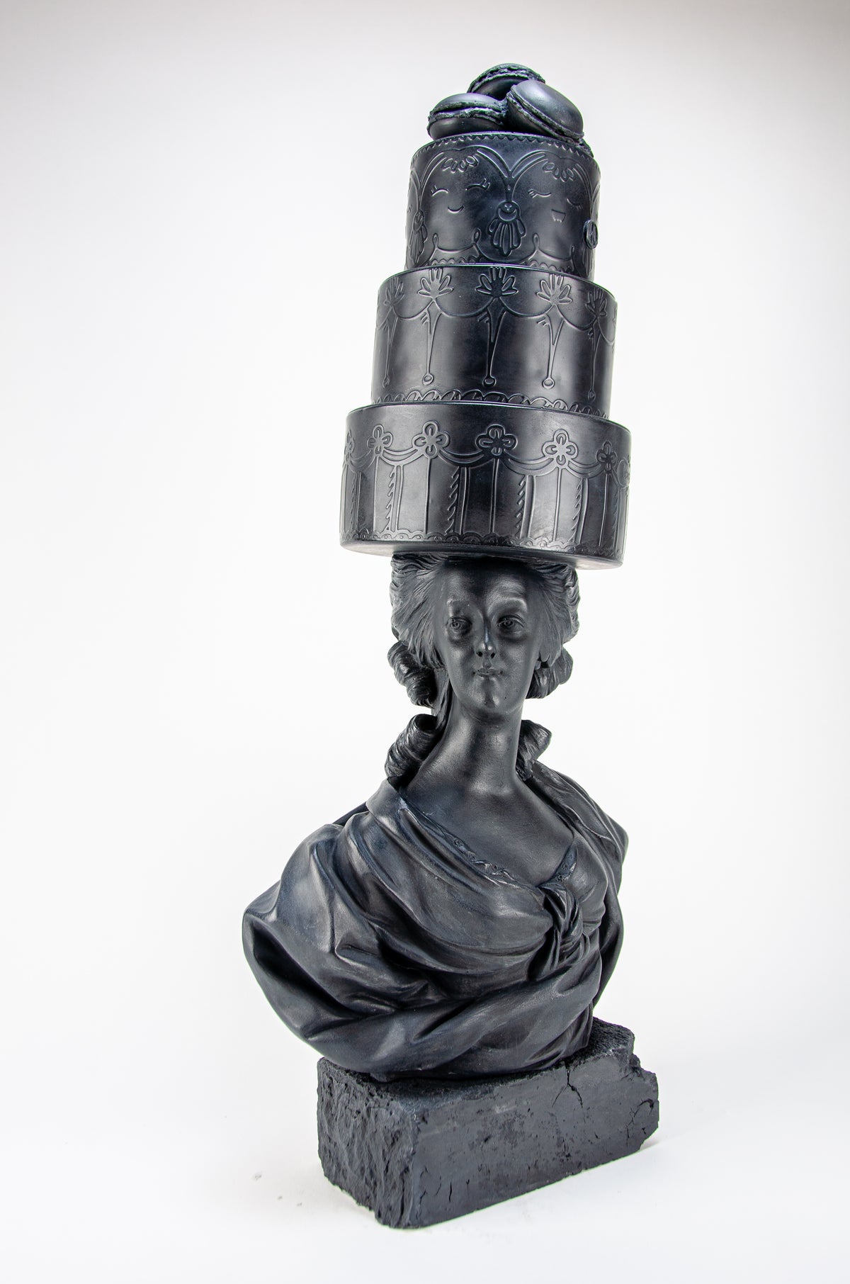 Tall dark charcoal sculpture of Marie Antoinette staring straight ahead wearing a giant three-layered cake with charcoal Macrons on top of her head.  