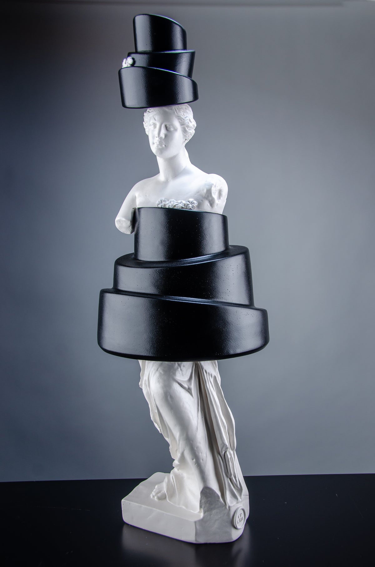 Tall white sculpture of Venus de Milo wearing a black cake that covers her body and a cake as a hat. The artful sculpture has small white flowers on top of the two cakes.