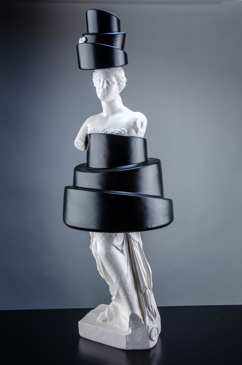 Tall white sculpture of Venus de Milo wearing a black cake that covers her body and a cake as a hat. The artful sculpture has small white flowers on top of the two cakes.