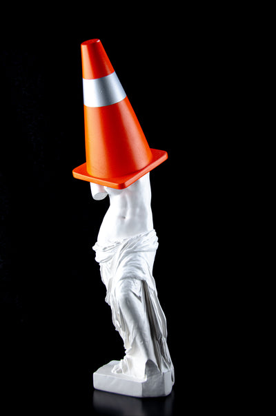 Tall white Venus de Milo sculpture with a large orange and silver traffic cone covering her head and part of her breasts. 