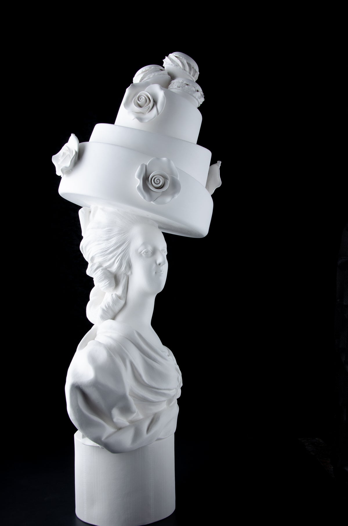 Tall bright white sculpture of Marie Antoinette looking facing forward wearing a large whimsical cake with roses and Macron cookies on her head. Dark black background.