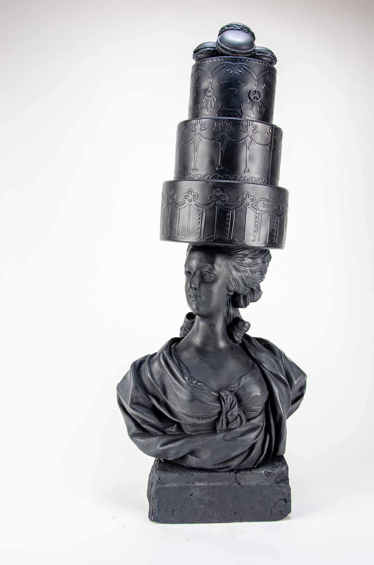 Tall dark charcoal sculpture of Marie Antoinette wearing a giant three-layered cake with charcoal Macrons on top of her head.