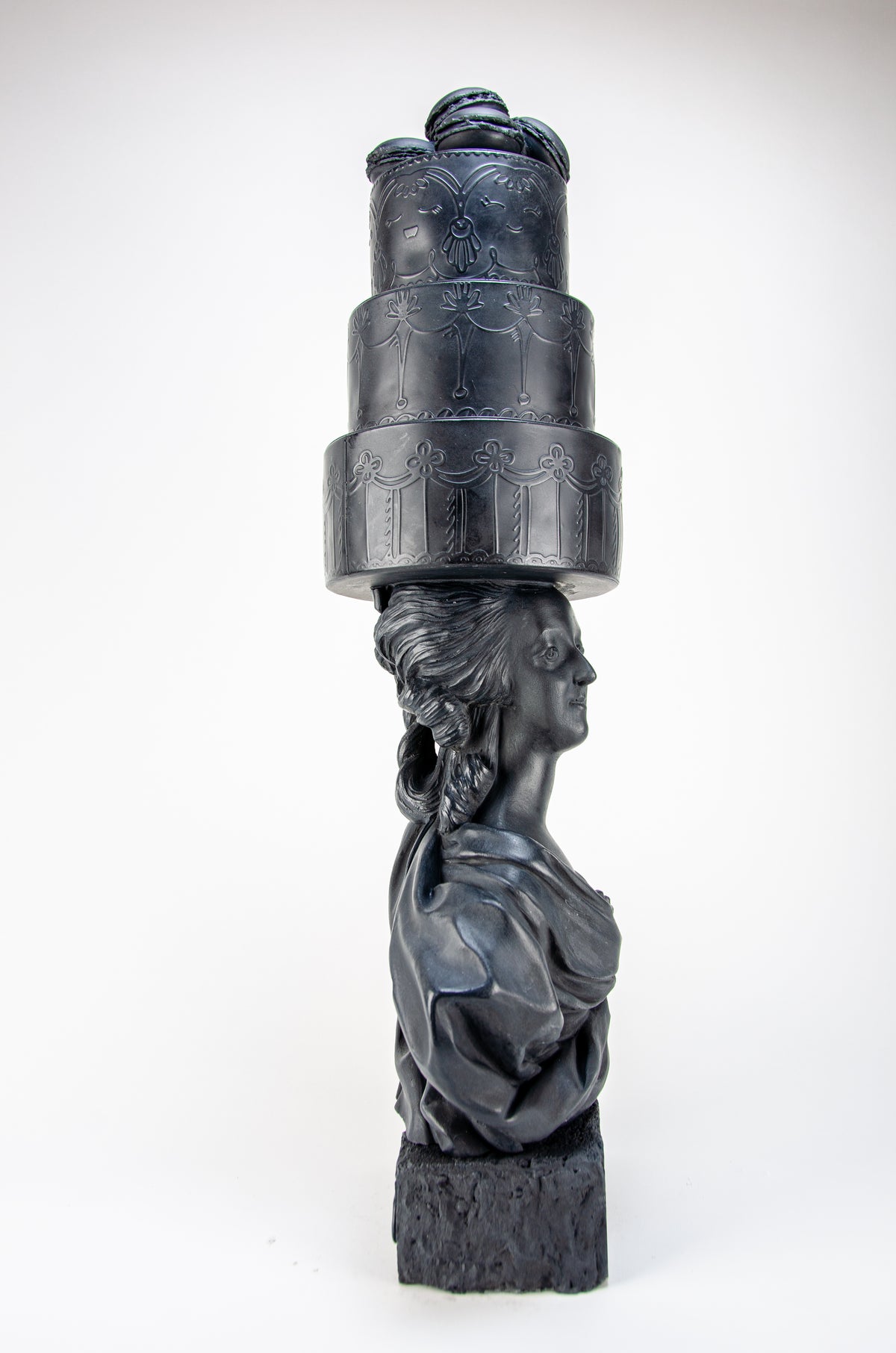 Tall dark charcoal sculpture of Marie Antoinette staring off into the distance wearing a giant three-layered cake with charcoal Macrons on top of her head.  