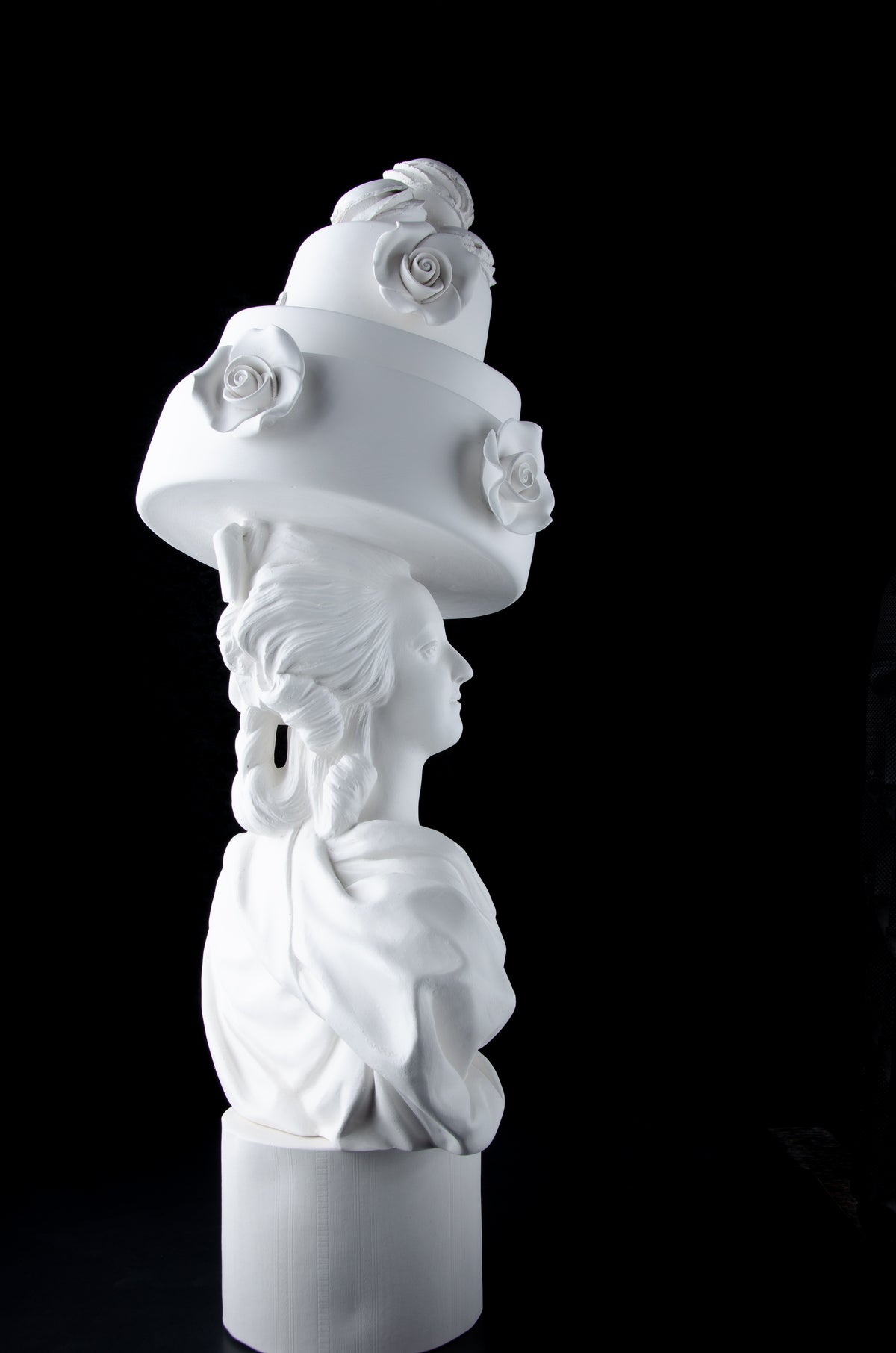 Tall bright white sculpture of Marie Antoinette staring off to the right wearing a large whimsical cake with roses and Macron cookies on her head. Dark black background.