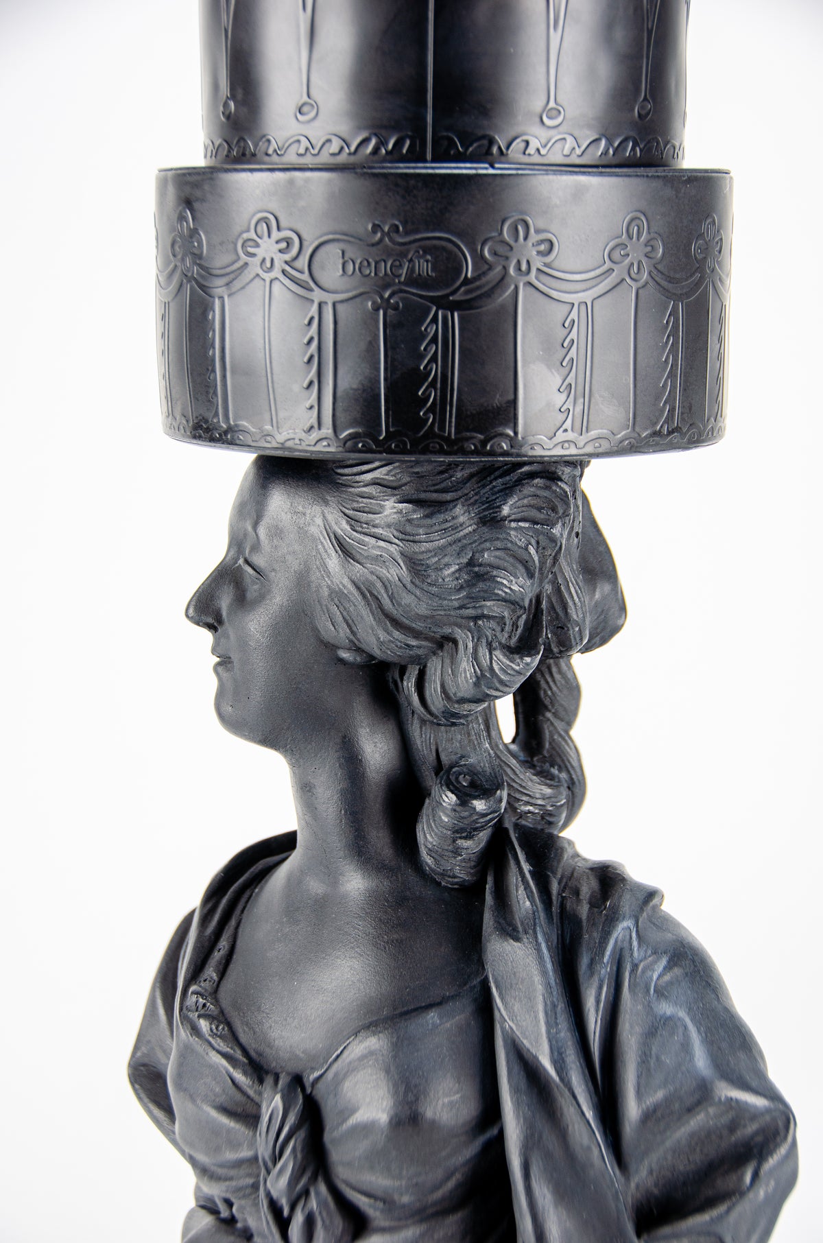 Detailed view of tall dark charcoal sculpture of Marie Antoinette looking to her left wearing a giant three-layered cake with charcoal Macrons on top of her head.