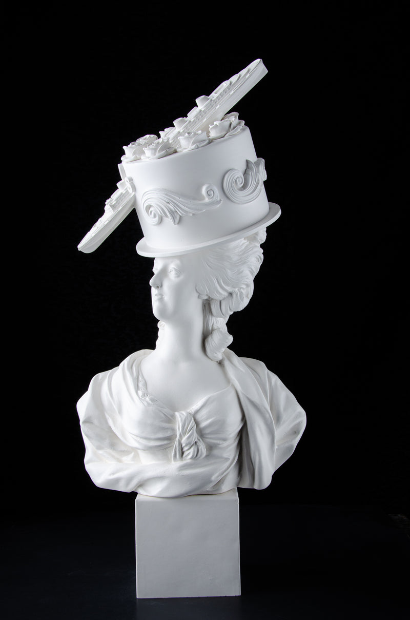 White sculpture bust of Marie Antoinette looking to the side wearing a large frosted with roses and scrolls cake with a battleship stuck in the side of the cake coming out the front on her head. Dark black background.