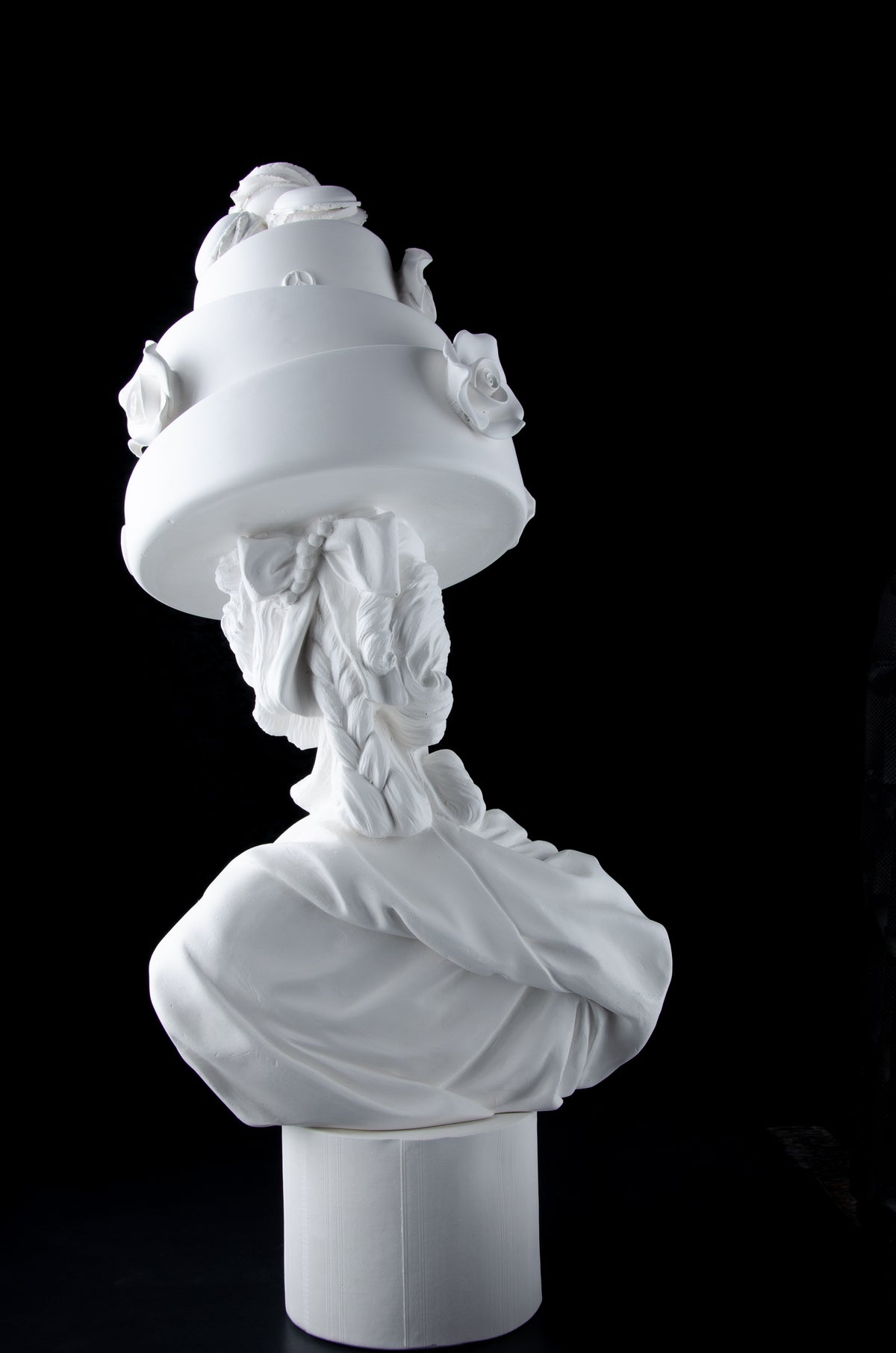 Tall bright white sculpture of Marie Antoinette viewed from the back wearing a large whimsical cake with roses and Macron cookies on her head. Dark black background.