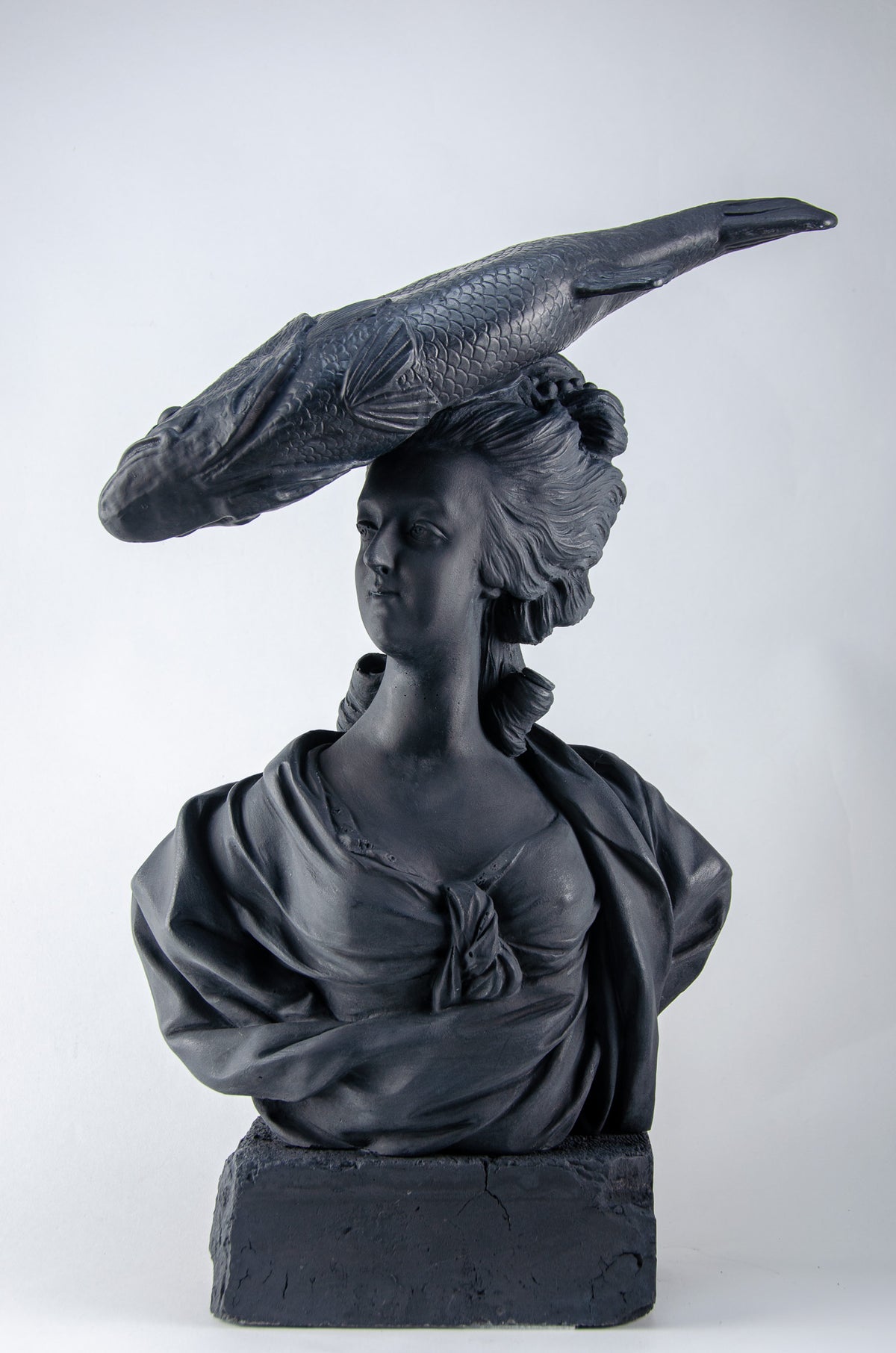 Deep charcoal bust sculpture of Marie Antoinette wearing a large fish on her head as a hat, she sits atop a brick of the same deep charcoal color.