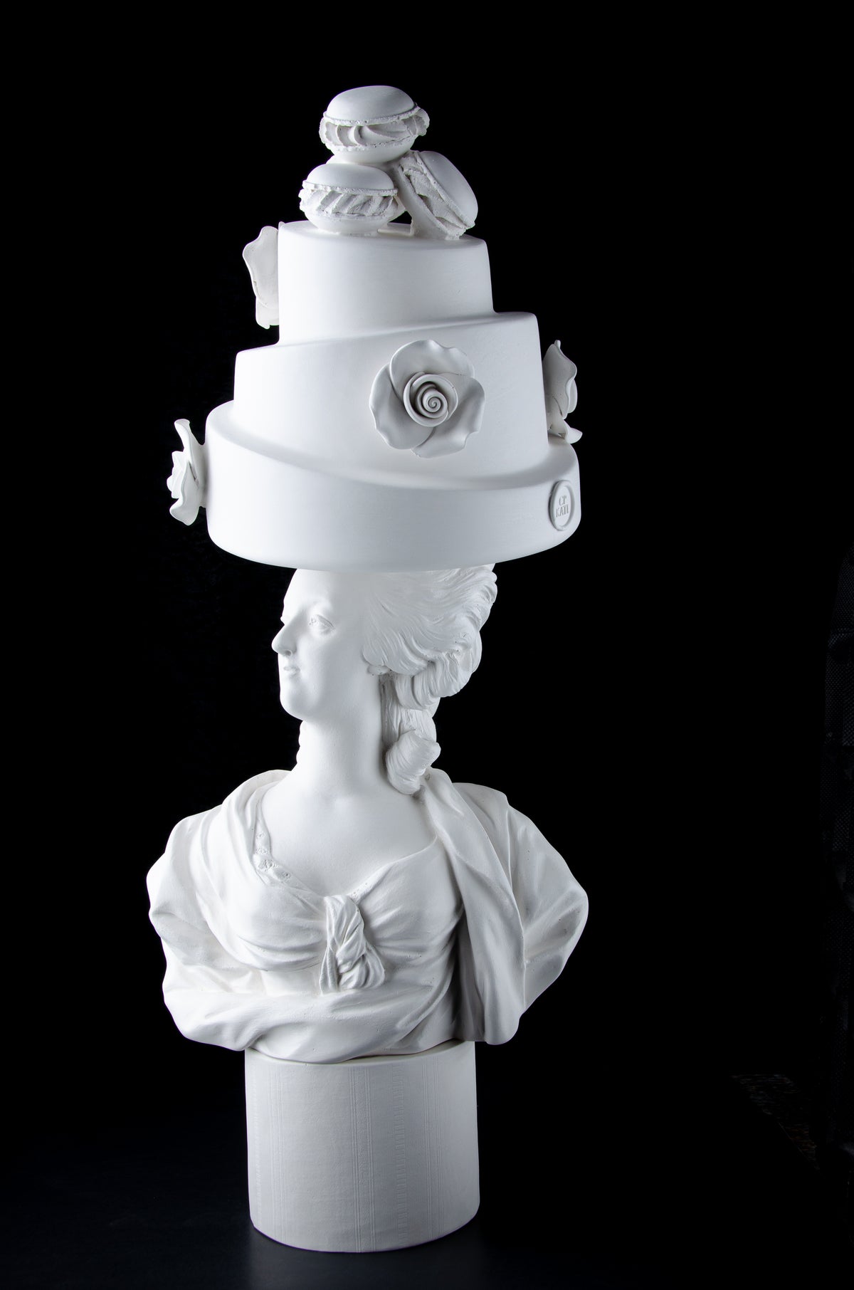 Tall bright white sculpture of Marie Antoinette looking to the side wearing a large whimsical cake with roses and Macron cookies on her head. Dark black background.