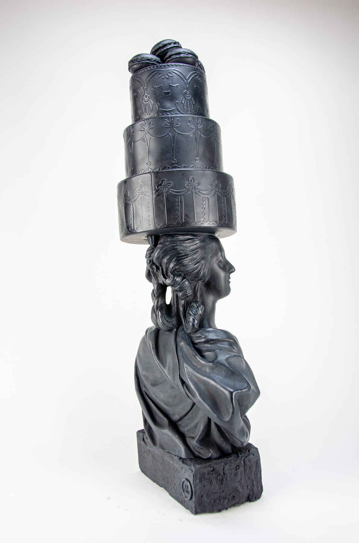Tall dark charcoal sculpture of Marie Antoinette staring off into the distance from her right side wearing a giant three-layered cake with charcoal Macrons on top of her head.  