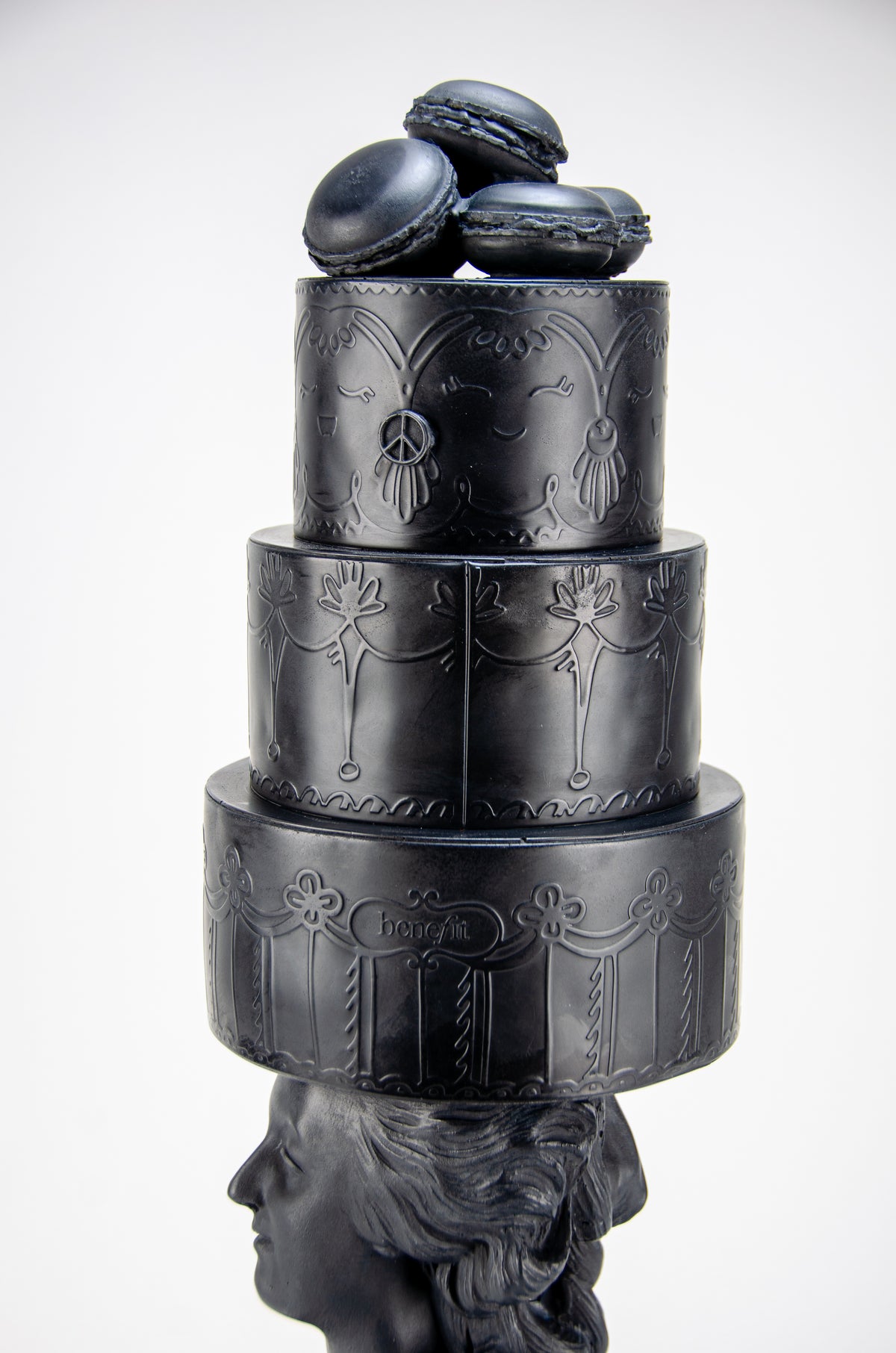 Detailed view of tall dark charcoal sculpture of Marie Antoinette staring off to her left wearing a giant three-layered cake with charcoal Macrons on top of her head.