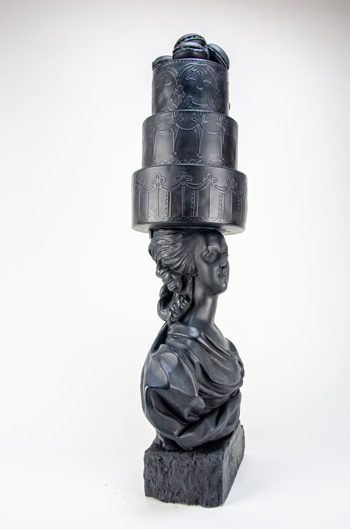 Tall dark charcoal sculpture of Marie Antoinette looking to her right wearing a giant three-layered cake with charcoal Macrons on top of her head.  