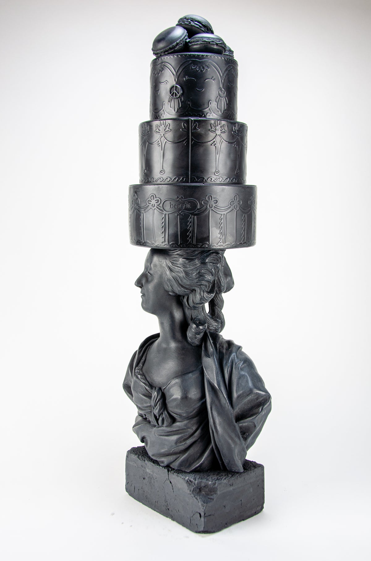 Tall dark charcoal sculpture of Marie Antoinette looking to her left wearing a giant three-layered cake with charcoal Macrons on top of her head.
