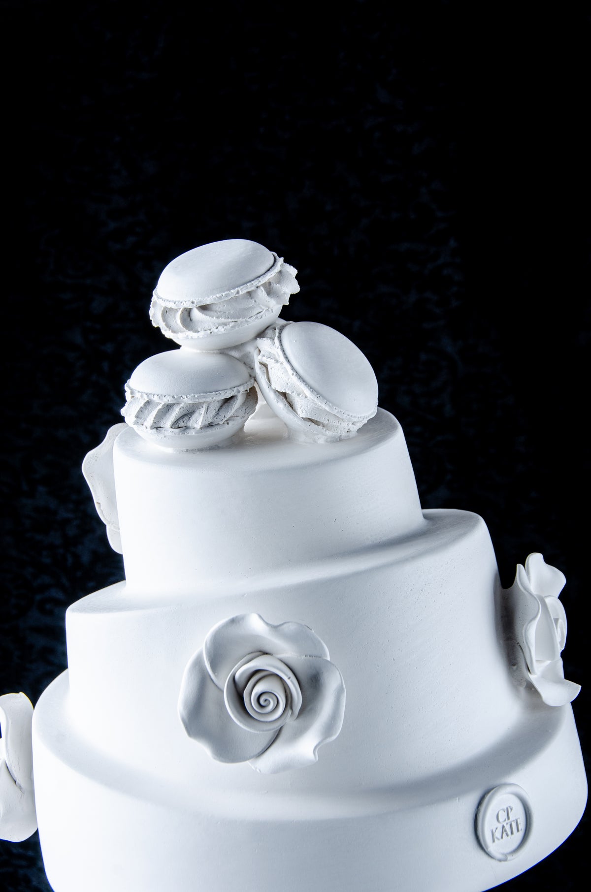 Detailed picture of a sculpture bust of Marie Antoinette looking to the side wearing a large whimsical cake with roses and Macron cookies on her head. Dark black background.