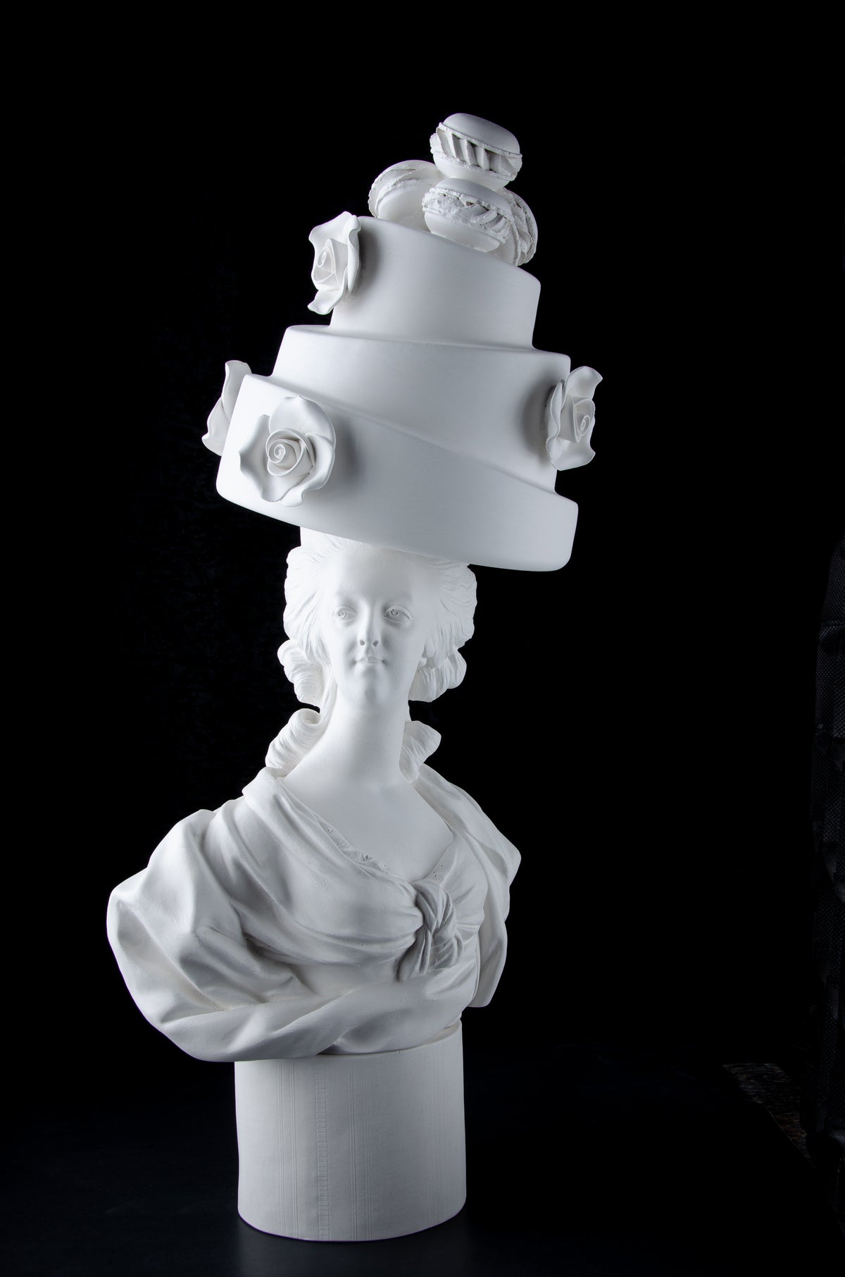 White sculpture bust of Marie Antoinette looking forward and wearing a large whimsical cake with roses and Macron cookies on her head. Dark black background.