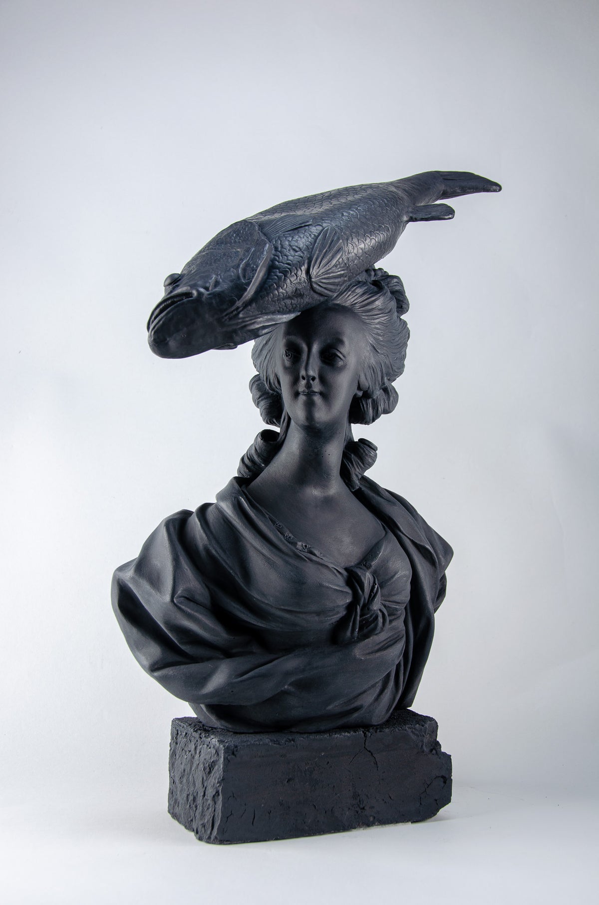Deep charcoal bust sculpture of Marie Antoinette staring straight ahead wearing a large fish on her head as a hat, she sits atop a brick of the same deep charcoal color.