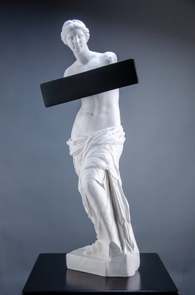 Tall white sculpture of Venus de Milo with a black band emblazoned across her chest as if being blocked or blurred by social media. The artful sculpture sits on top of a black plinth.