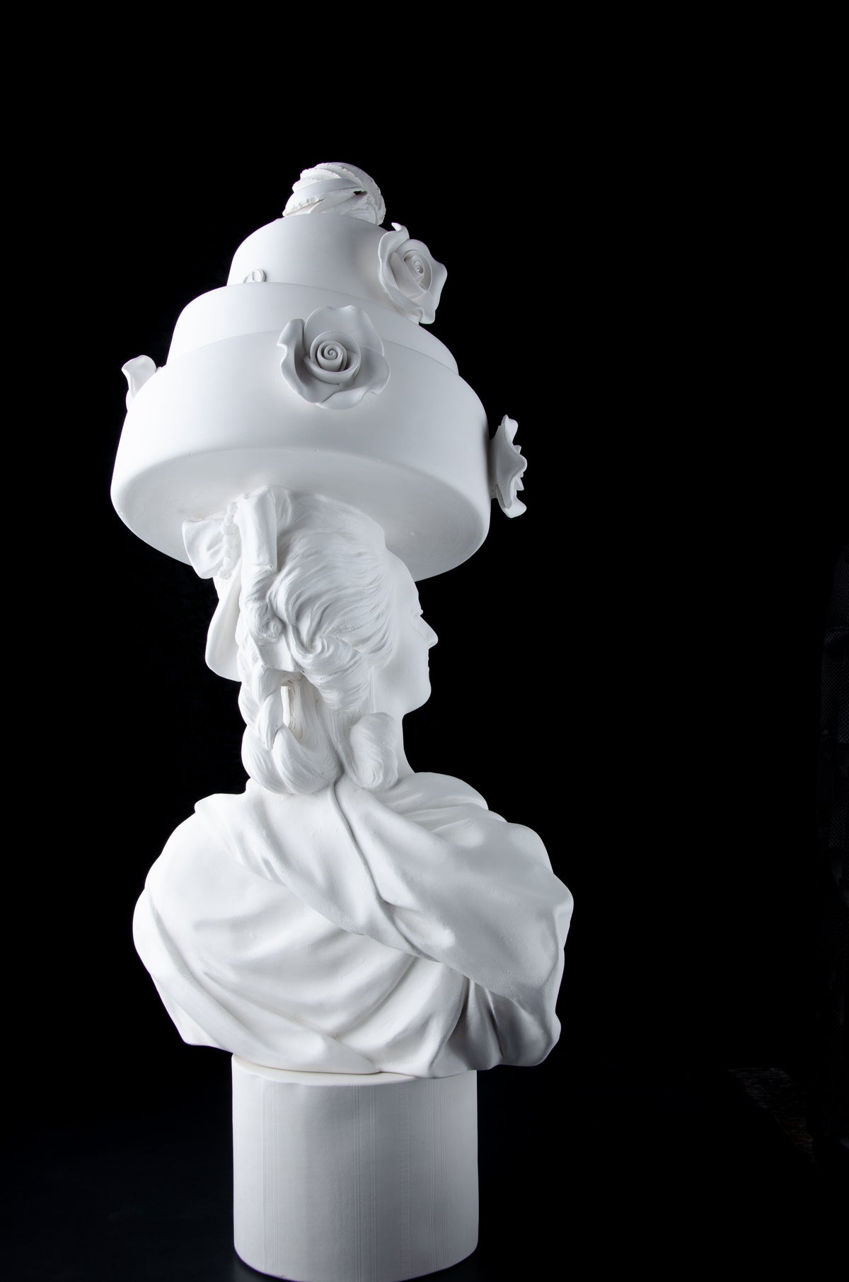 Tall bright white sculpture of Marie Antoinette looking to her right wearing a large whimsical cake with roses and Macron cookies on her head. Dark black background.