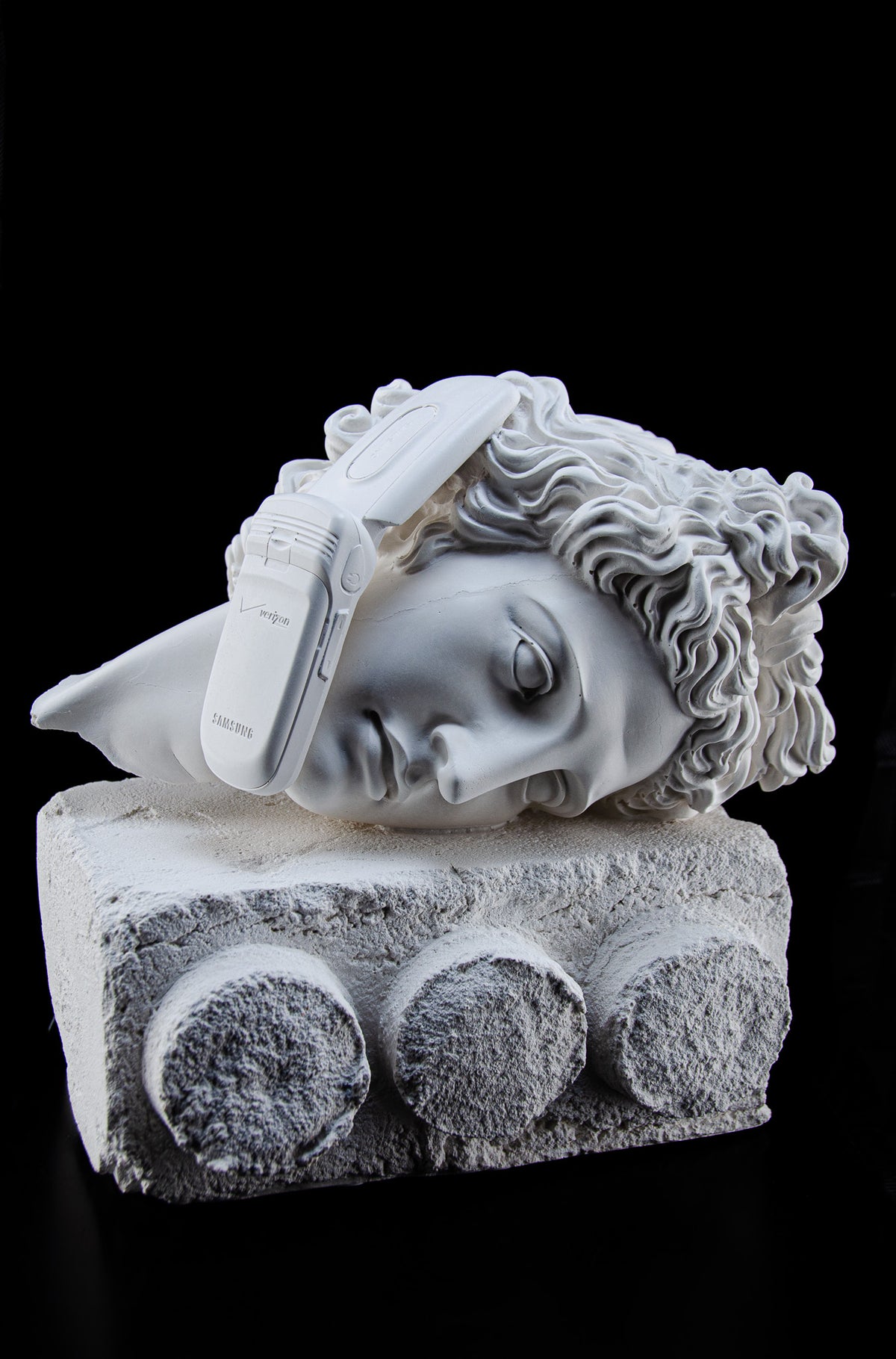 White sculpture of a broken bust of Apollo Belvedere's listening to an old-school flip cell phone. He sits atop a white plinth in the shape of an old-world Lego that looks as if it was carved from Stone.